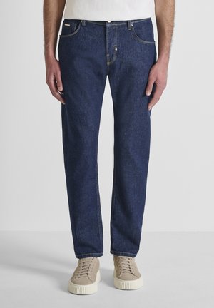 Man draagt donkere blauwe rechtgesneden jeans en beige sneakers, staand tegen een effen achtergrond, handen ontspannen langs het lichaam.