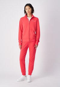 Sudadera roja con cremallera y joggers a juego, hechos de un tejido suave. Presenta un logo en el pecho y puños elásticos. Combinado con zapatillas deportivas blancas.