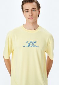 Camiseta de algodón amarilla de manga corta presenta una impresión gráfica azul de copas de cóctel y el texto "BEBIDAS DELICIOSAS" debajo.