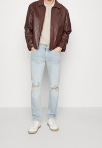 Chaqueta de cuero marrón sobre una camisa beige, combinada con vaqueros desgastados azul claro y zapatillas blancas con suela color canela.