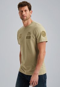 Beige bomulds t-shirt med rund hals, udstyret med præget tekst og et cirkulært emblem på ærmet. Designet til en tætsiddende pasform.