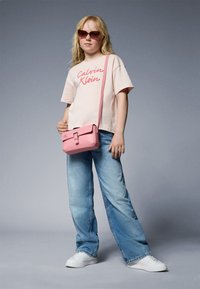 T-shirt cropped rosa chiaro con scritta "Calvin Klein", jeans a gamba larga blu, borsa a spalla rosa e sneakers bianche. Gli accessori includono occhiali da sole chiari.