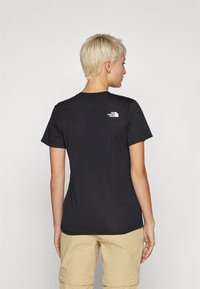 Oseba z kratkimi blond lasmi, oblečena v črno majico The North Face in bež kratke hlače, gledana s hrbta proti enobarvni beli ozadju.