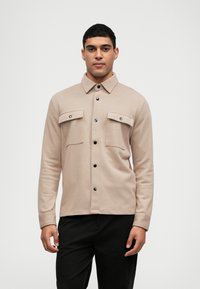Camicia beige con bottoni a pressione, colletto, due tasche sul petto e bottoni neri a scatto. Realizzata in un tessuto texturizzato. Indossata sopra pantaloni scuri.