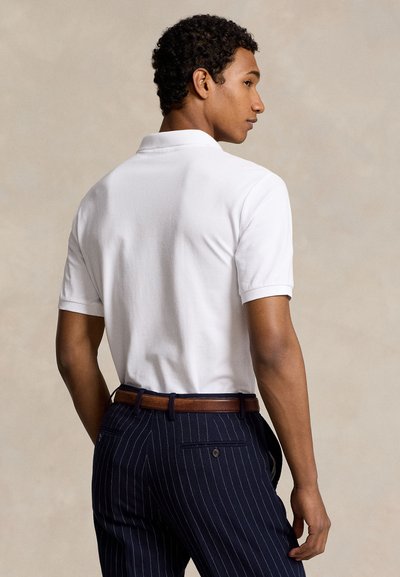 Muž v bielej krátkosleevej polo košeli zastrčenej do námorníckych pásikavých nohavíc s hnedým opaskom, stojaci chrbtom a mierne otočený hlavou doprava.