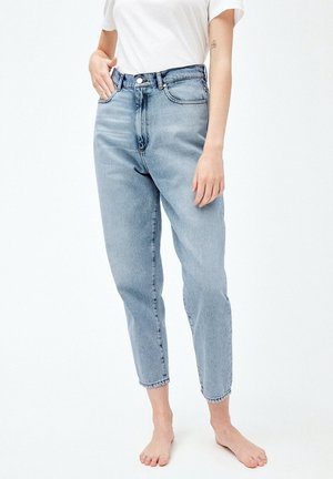 Femme portant un jean taille haute bleu clair et un t-shirt blanc, debout pieds nus avec une main dans la poche et un tatouage visible sur le bras.