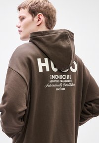 Rudos spalvos džemperis su dideliu baltu „HUGO“ logotipu nugaroje, taip pat su auksinėmis detalėmis ir prekės ženklo užrašu. Pagamintas iš tekstūruotos medžiagos su gobtuvu.