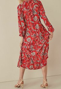 Robe longue rouge à manches longues avec des motifs floraux et feuillus, cintrée à la taille, jupe fluide et texture de tissu douce. Présente des sandales à bride de cheville.