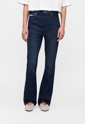 Personne portant un jean bootcut bleu foncé et des chaussures beige clair sans lacets, debout devant un fond blanc uni.