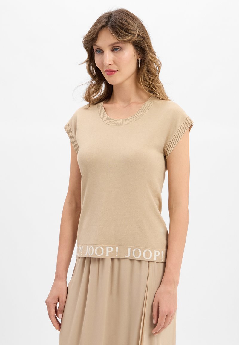 Frau mit welligem Haar, die ein beigefarbenes ärmelloses Top mit dem Logo "JOOP!" am Saum und einen passenden beigefarbenen Rock trägt, steht vor einem weißen Hintergrund.