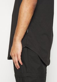 Černé tričko vyrobené z měkké bavlny, se mírně zakřiveným lemem a štítkem na postranním švu. Nosené přes tmavé cargo kalhoty.