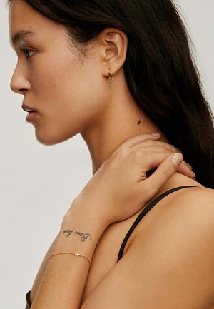 Pendientes de aro dorados con un detalle de ancla, una delicada pulsera dorada con un acento de cruz y una textura de piel suave contra una blusa oscura.