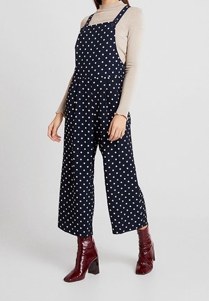 Kvinde iført marineblå vidbenede overalls med prikker, beige langærmet top og mørkerøde ankelstøvler med hæl på neutral baggrund.