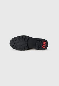 Semelle de chaussure en caoutchouc noir avec un motif de grip texturé et un logo "HU" rouge près du talon, mettant en avant un design robuste pour une meilleure adhérence.