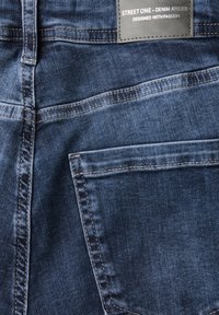 Jeans en denim bleu foncé avec une texture lisse, des coutures visibles et une poche arrière. L'étiquette indique "STREET ONE - DENIM ATELIER."