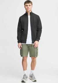 Sweatshirt zippé noir, t-shirt blanc en dessous, shorts cargo verts, chaussettes blanches, chaussures de sport grises. Design simple et fonctionnel.