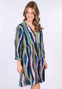 Mehrfarbiges Kleid mit V-Ausschnitt, Dreiviertelärmeln und ausgestellter Silhouette. Der Stoff zeigt Zickzackmuster in Grün, Blau und Lila.