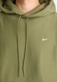 Nærbillede af en olivenfarvet hoodie med snørebånd og et lille hvidt Nike swoosh-logo på brystet.
