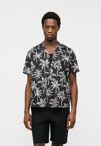 Νεαρός άνδρας που φοράει μαύρο πουκάμισο με κοντά μανίκια, κουμπωτό, με λευκή εκτύπωση φοίνικα και μαύρο σορτς, στέκεται μπροστά από ανοιχτό φόντο.