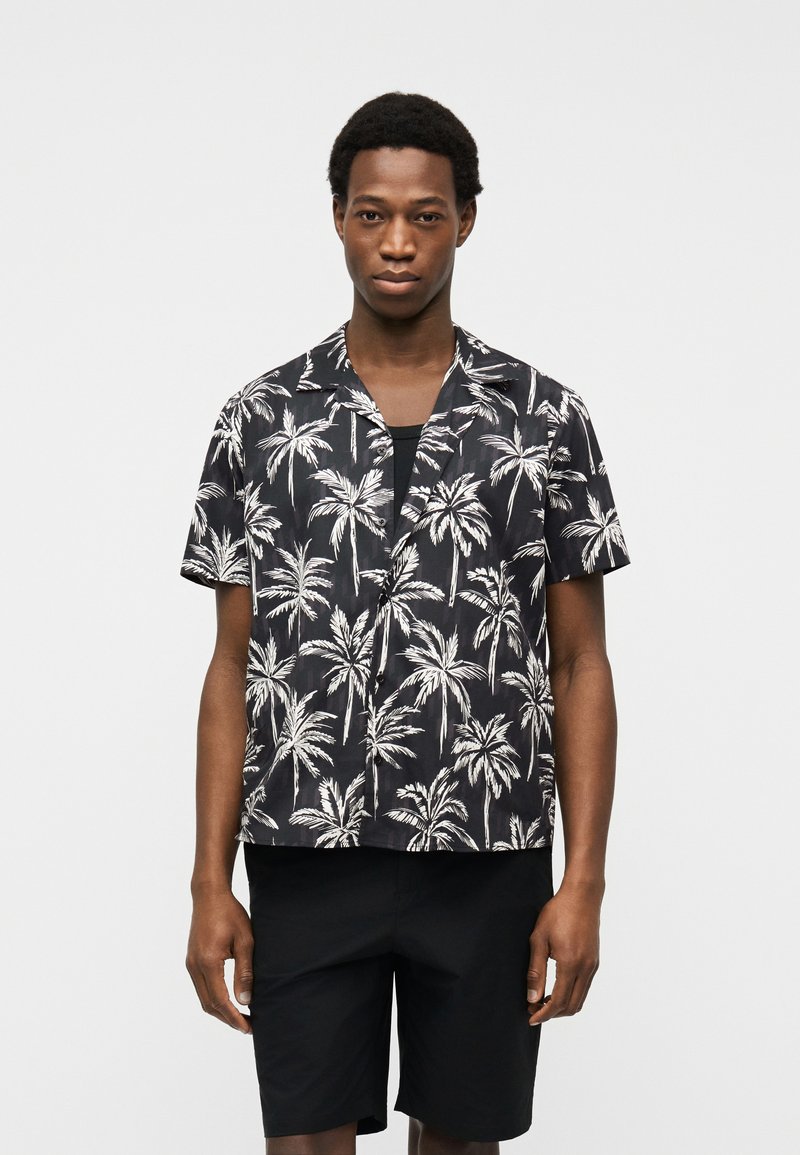 Νεαρός άνδρας που φοράει μαύρο πουκάμισο με κοντά μανίκια, κουμπωτό, με λευκή εκτύπωση φοίνικα και μαύρο σορτς, στέκεται μπροστά από ανοιχτό φόντο.