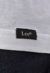 Klein zwart stoffen label met witte tekst "Lee®" genaaid op de zoom van een wit kledingstuk boven donker denimstof.