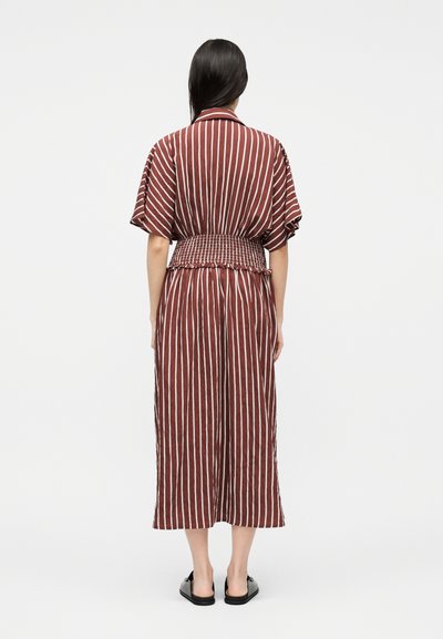 Γυναίκα που φορά ένα μίντι φόρεμα με κάθετες καφέ και λευκές ρίγες, κοντά μανίκια και φαρδιά ελαστική μέση, όρθια με την πλάτη προς το μέρος μας, σε απλό φόντο.