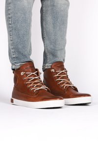 Scarpe alte in pelle marrone con lacci beige e suola in gomma bianca. Presentano un logo circolare sul lato. Indossate con jeans azzurri chiari.