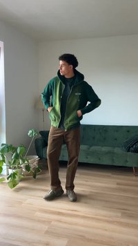 Vihreä gradienttinen vetoketjuhuppari tumman paidan yllä, ruskeat corduroyhousut ja harmaat lenkkarit, seisoo hyvin valaistussa huoneessa, jossa on kasveja.