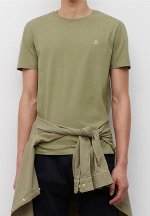 Olijfgroene katoenen T-shirt met ronde hals en korte mouwen, met een klein wit logo op de borst, gecombineerd met een lichtbeige overhemd dat om de taille is gebonden.