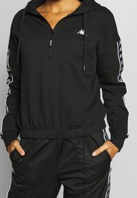 Sweat-shirt à capuche noir avec fermeture éclair, taille élastique et poches latérales ; présente un logo blanc et un ruban à motifs sur les manches.