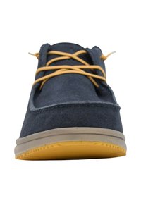 Zapato de ante azul oscuro con cordones amarillos, suela marrón ligeramente elevada y detalles mínimos de costura. Punta redondeada y diseño de perfil bajo.