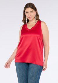 Top senza maniche in raso rosso con scollatura a V, forma fluida e texture liscia. Indossato con pantaloni a vita alta blu con righe verticali.