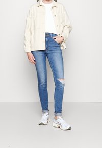 Chaqueta beige de botón sobre una camisa blanca, combinada con jeans ajustados rasgados azul y zapatillas deportivas blancas con acentos grises.
