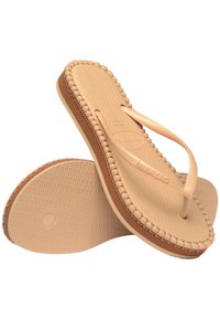 Tongs tan avec des fines brides, semelles texturées et bords cousus. Matériau en caoutchouc lisse et logo de marque notable sur la semelle.