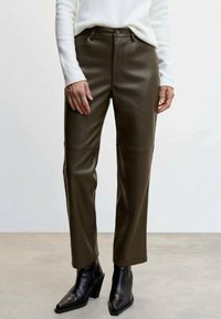 Olivgrüne Kunstlederhose mit geradem Bein, Vordertaschen und Metallknopfverschluss, kombiniert mit schwarzen Stiefeletten mit Absatz.