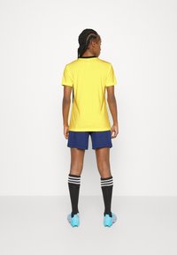 Geltona sportinė marškinė su juodu apykakle, tamsiai mėlynės šortai, juodos kelnės iki kelių su baltais dryžiais ir akvamarino spalvos futbolo batai.