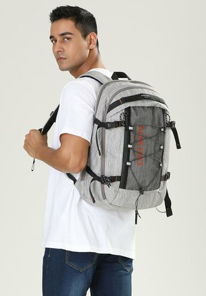 Man draagt wit T-shirt en spijkerbroek, en draagt een grote grijze Aoking-rugzak met meerdere vakken en zijriemen.