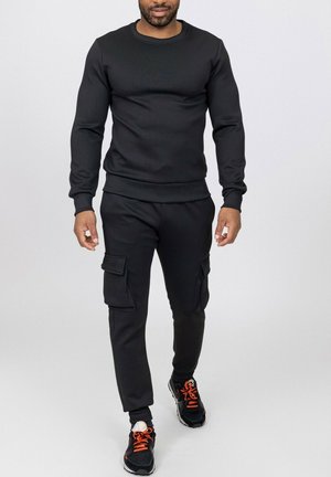 Homme portant un sweat-shirt noir à manches longues, un pantalon cargo noir avec des poches et des baskets noires avec des lacets orange, debout sur un fond uni.