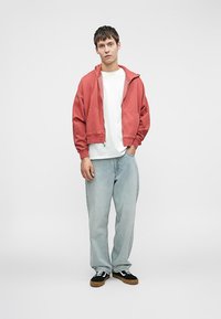 Κόκκινη ζακέτα με φερμουάρ πάνω από ένα λευκό T-shirt, συνδυασμένη με ανοιχτό μπλε τζην και μαύρα αθλητικά παπούτσια με καφέ σόλες. Λείος υφασμάτινος ιστός σε όλη την επιφάνεια.