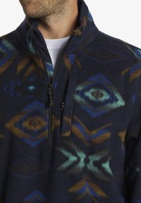 Fleece-Pullover in Dunkelblau mit geometrischen Mustern in Türkis, Braun und Gold. Verfügt über einen Reißverschluss und eine kleine Brusttasche. Weiche Textur.