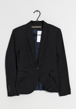 Blazer noir à un bouton avec revers crantés, poches à rabat et une étiquette blanche accrochée à l'intérieur, présenté sur un cintre noir sur un fond blanc.