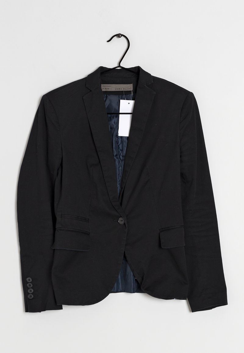 Blazer noir à un bouton avec revers crantés, poches à rabat et une étiquette blanche accrochée à l'intérieur, présenté sur un cintre noir sur un fond blanc.