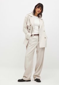 Bežinė dvigubo užsegimo tranšėja su dideliais mygtukais, derinama su balta prigludusia marškinėliais ir kreminėmis plačiomis kelnėmis. Rudas diržas ir juodi batai.