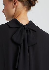 Blouse noire avec un col rond et un nœud attaché à l'arrière. Tissu lisse et fermeture éclair invisible au niveau du cou.