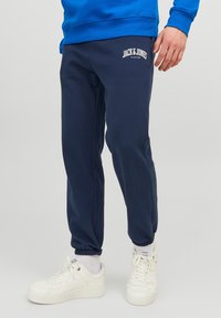 Navy blue sweatpants made of cotton blend, featuring a white logo on the left thigh and ribbed cuffs at the ankles. Worn with white sneakers.