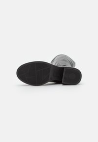 Botte noire allongée sur le côté montrant une semelle à motifs et un petit talon carré sur un fond blanc uni.