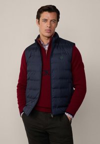Temno plavi quilted prsluk s visokim ovratnikom, uparen s burgundskim puloverom i kariranom košuljom. Sadrži zeleni amblem na prsluku.