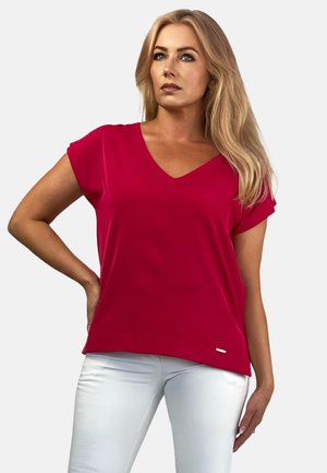 Czerwony t-shirt z krótkim rękawem, z dekoltem w serek, zaokrąglonym dołem i subtelnym rozcięciem po bokach, zestawiony z białymi spodniami. Materiał wydaje się gładki i lekki.