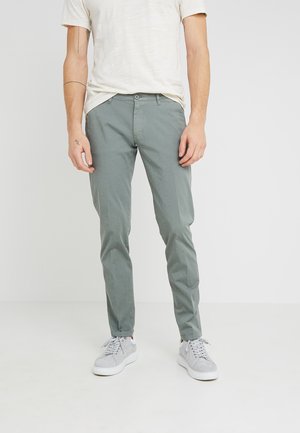 Pantalon en coton vert avec une coupe fuselée, comportant une fermeture à bouton à l'avant, des poches latérales et une texture lisse. Porté avec des baskets grises.