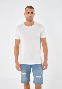 T-shirt bianca a maniche corte con scollatura rotonda e rifiniture a contrasto, abbinata a pantaloncini di jeans azzurri chiari con dettagli sfilacciati.